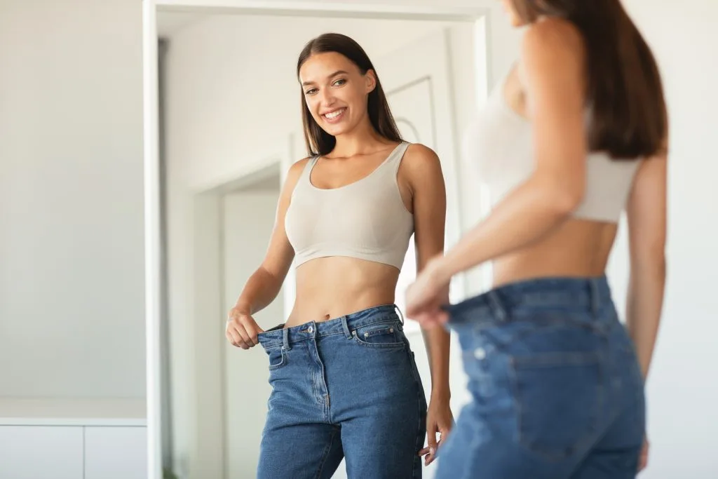 Woman demonstrating weight loss results by pinching loose jeans at the waist in front of a mirror
