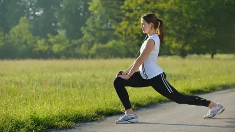 Woman stretching in a lunge position on a path with green grass and trees in background