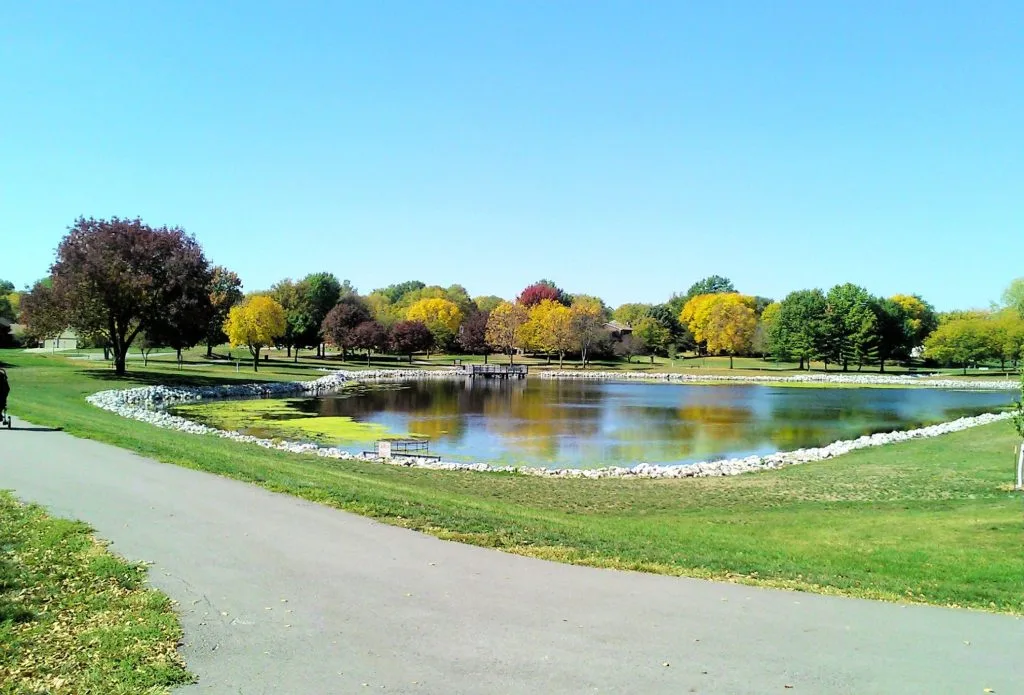 Park pathway beside pond with colorful autumn trees reflected in water and green lawn