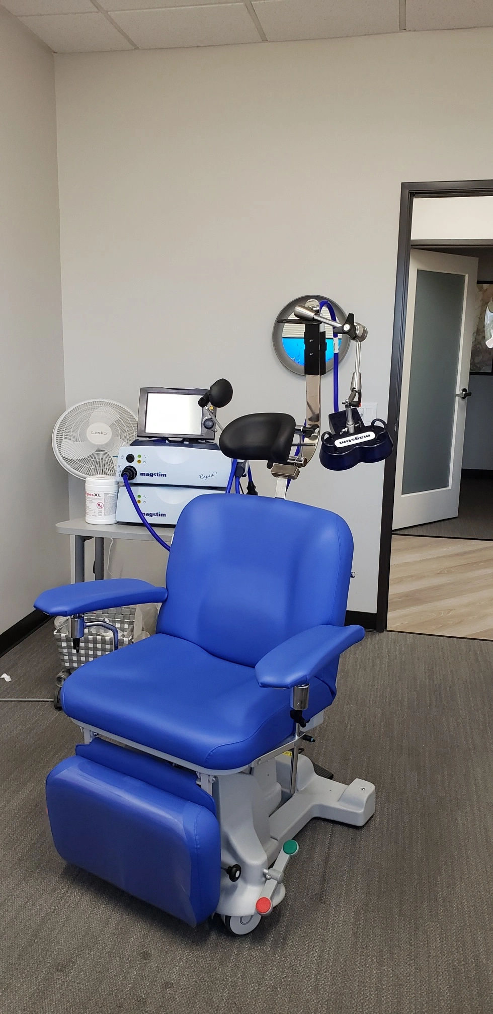 TMS therapy room with blue treatment chair, Magstim stimulation equipment, laptop, coil apparatus, and support furniture
