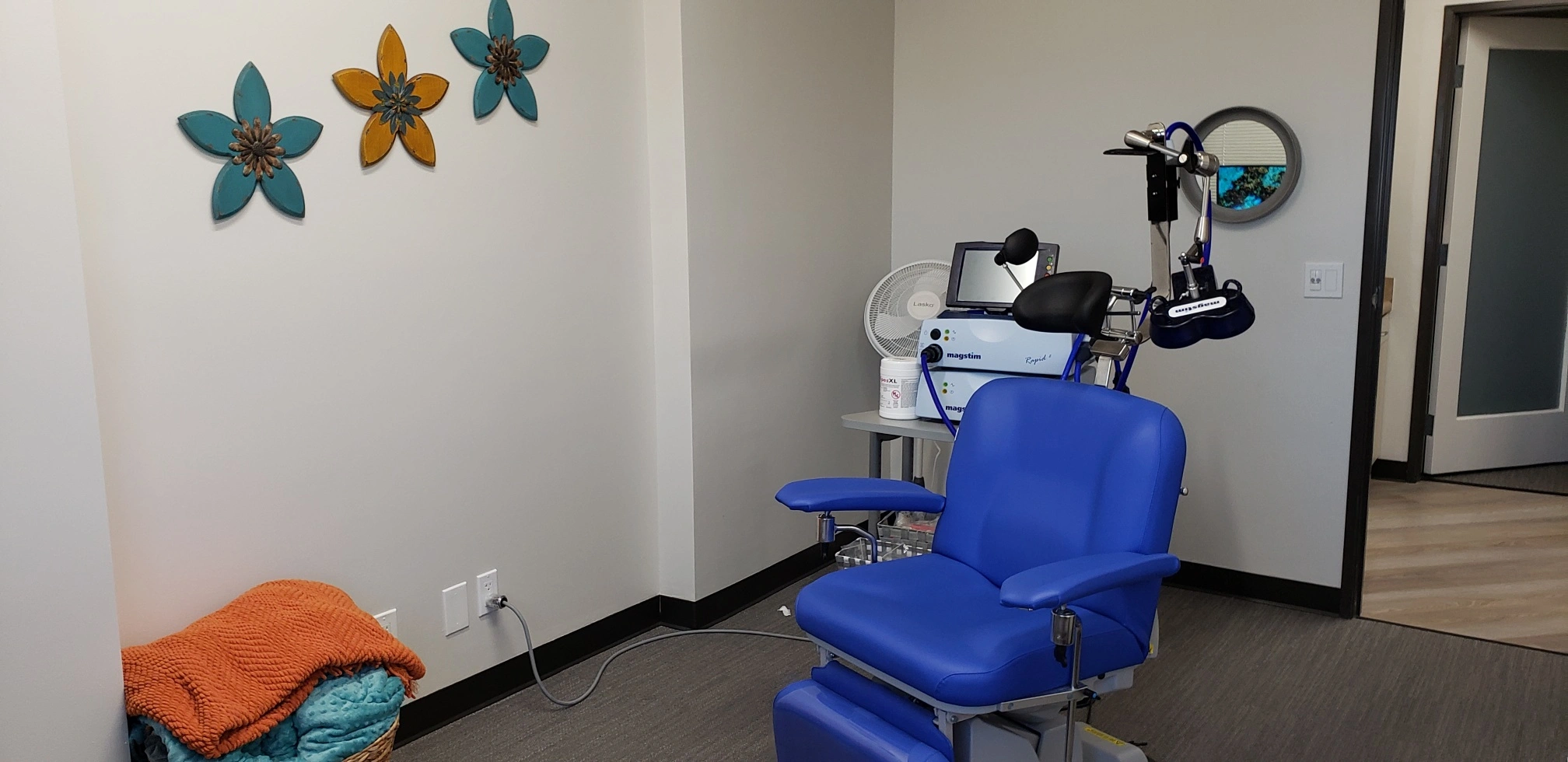 TMS treatment room with blue medical chair, Magstim device on white table, colorful flower wall decals, and equipment setup