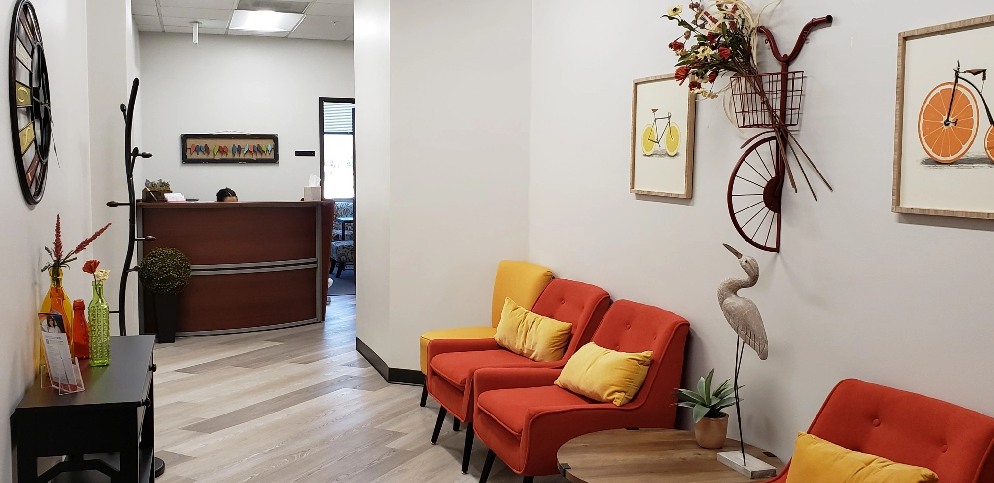 Reception area with dark wood desk, red and yellow cushioned seating, decorative wall art including bicycle and citrus artwork