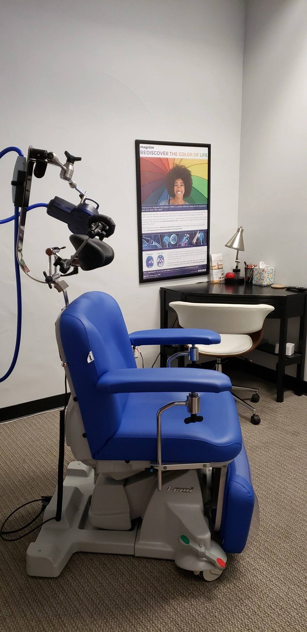 TMS treatment chair with mounted coil applicator and blue cables, poster reading 'Rediscover the Color of Life' on wall behind desk