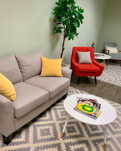 Waiting area with gray sofa, yellow pillow, red armchair, potted plant, and white coffee table with magazines