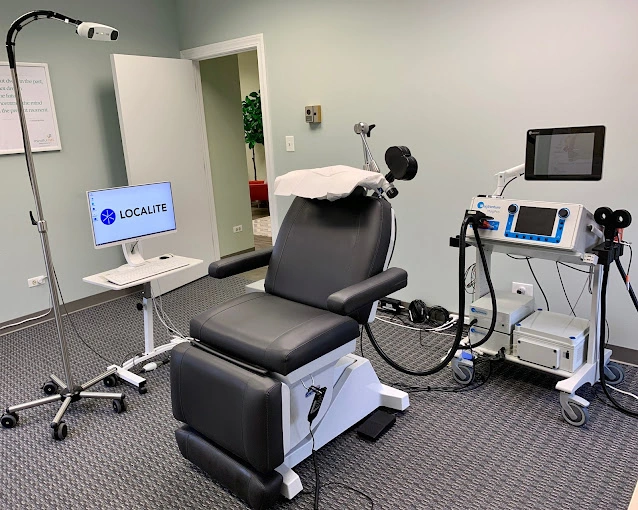 TMS treatment room with black patient chair, Localite monitor, overhead magnetic coil apparatus, and blue treatment device on cart