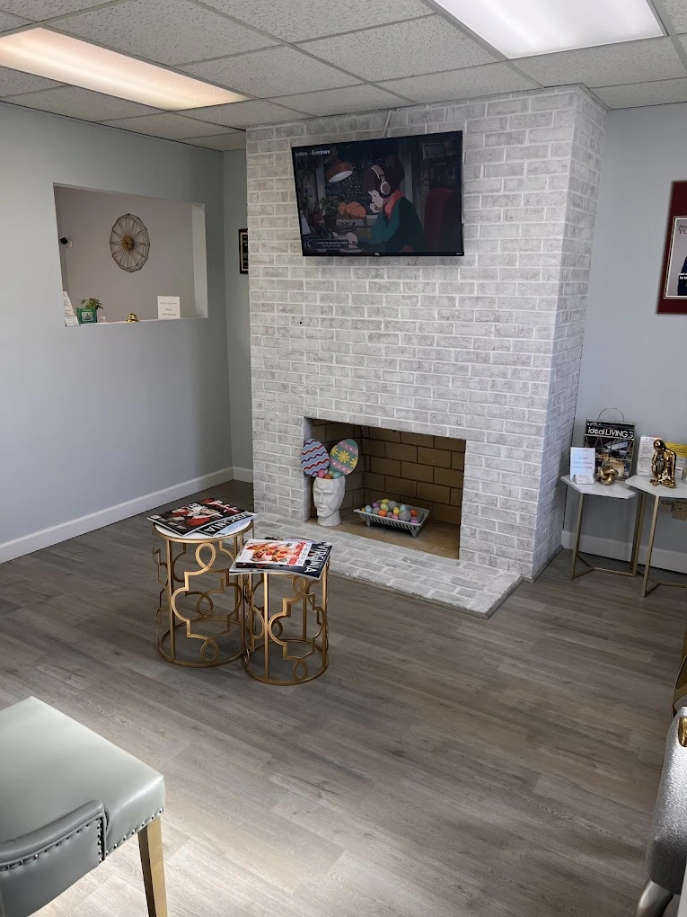 Medical office waiting room with white brick fireplace, TV mounted above, decorative side tables with magazines, and gold accent chair