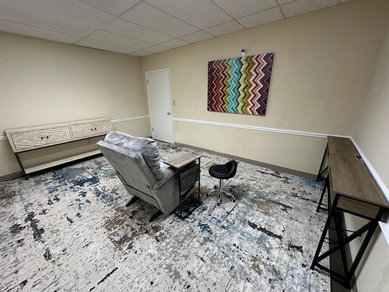 TMS treatment room with gray armchair, wooden benches, patterned wall hanging, and geometric floor rug
