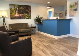 Waiting area with blue reception desk, gray sofa, leather chairs, landscape paintings, and desk lamp
