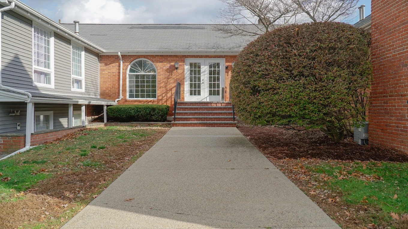 Brick building exterior with concrete walkway, white trim windows, and landscaped shrubs on both sides