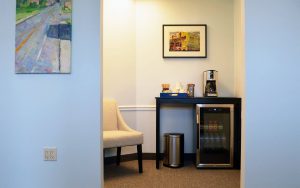 Bright clinic break room with cream chair, black desk, coffee maker, beverage cooler, and framed artwork on blue walls