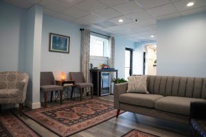 Modern clinic waiting area with gray upholstered seating, warm lighting, decorative rug, plants, and relaxation lamp