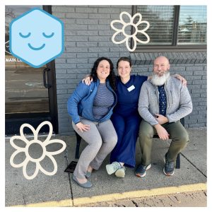 Three smiling staff members standing outside brick building entrance with decorative flower elements
