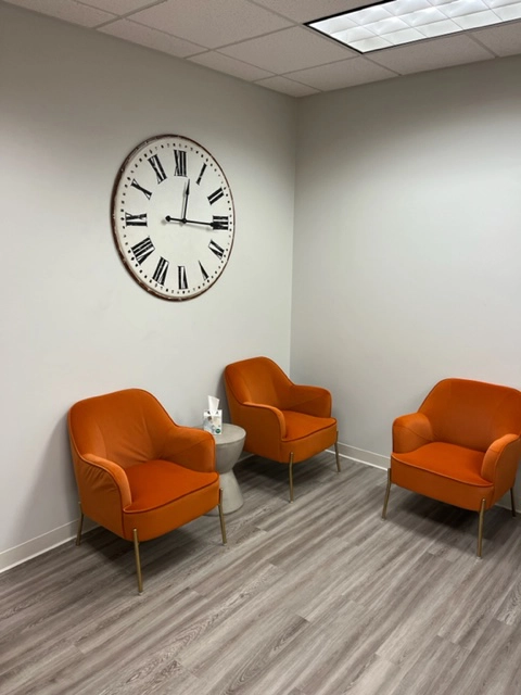 Clinical waiting area with three orange upholstered chairs, a small side table, and a wall clock