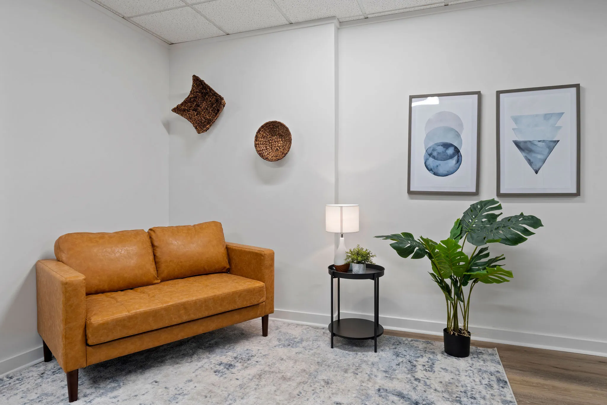 Therapy room with tan leather sofa, woven wall art, framed abstract prints, potted plant, and side table