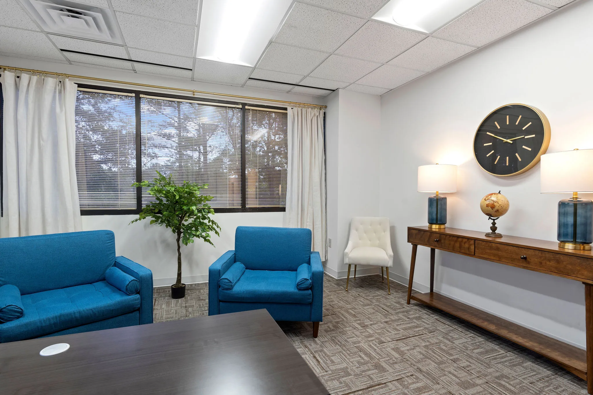 Clinical consultation room with blue sofa and armchair, wall clock, potted plant, and windows with curtains