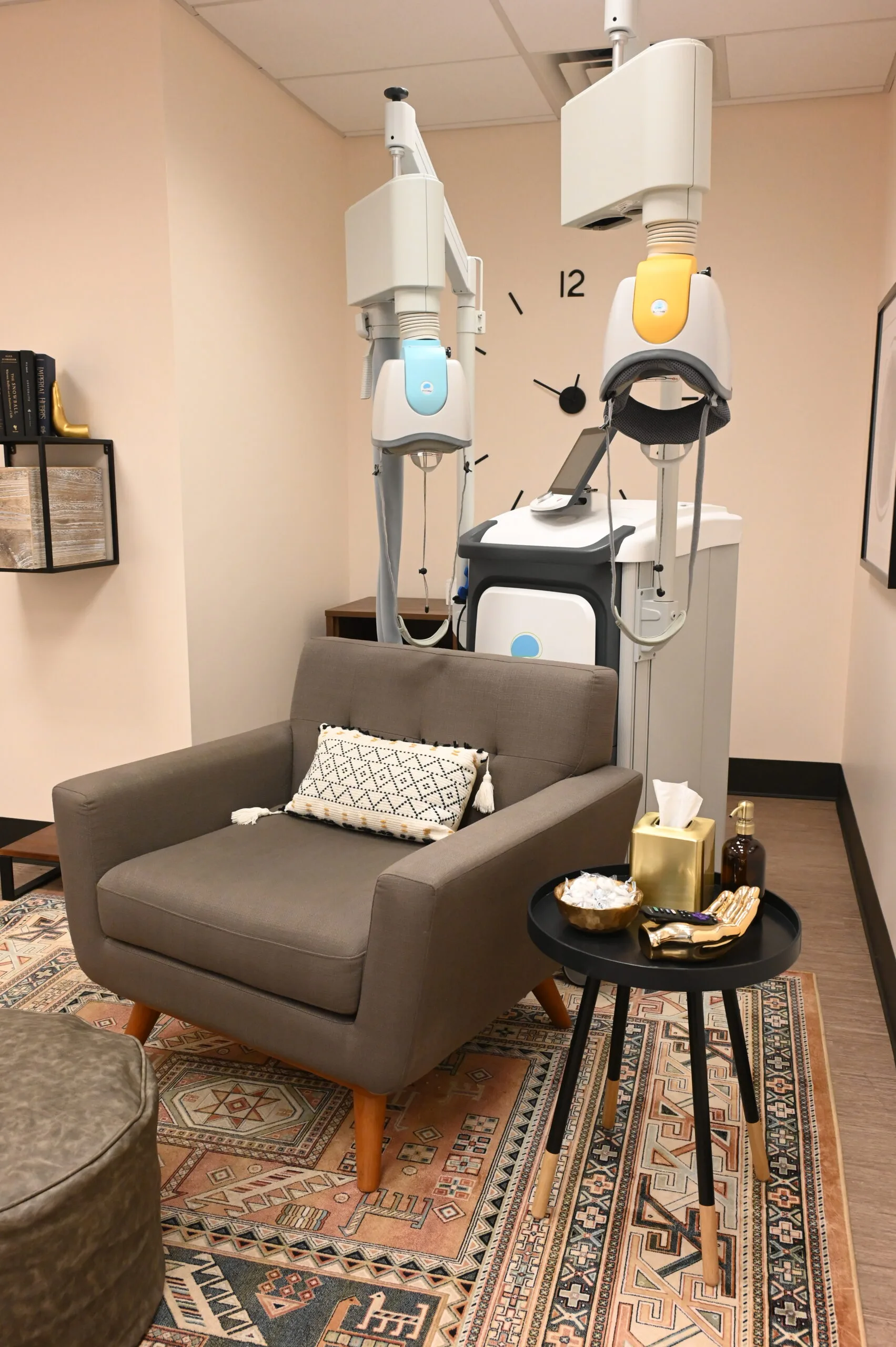 TMS therapy treatment room with gray armchair, overhead medical equipment with blue and yellow components, and black side table
