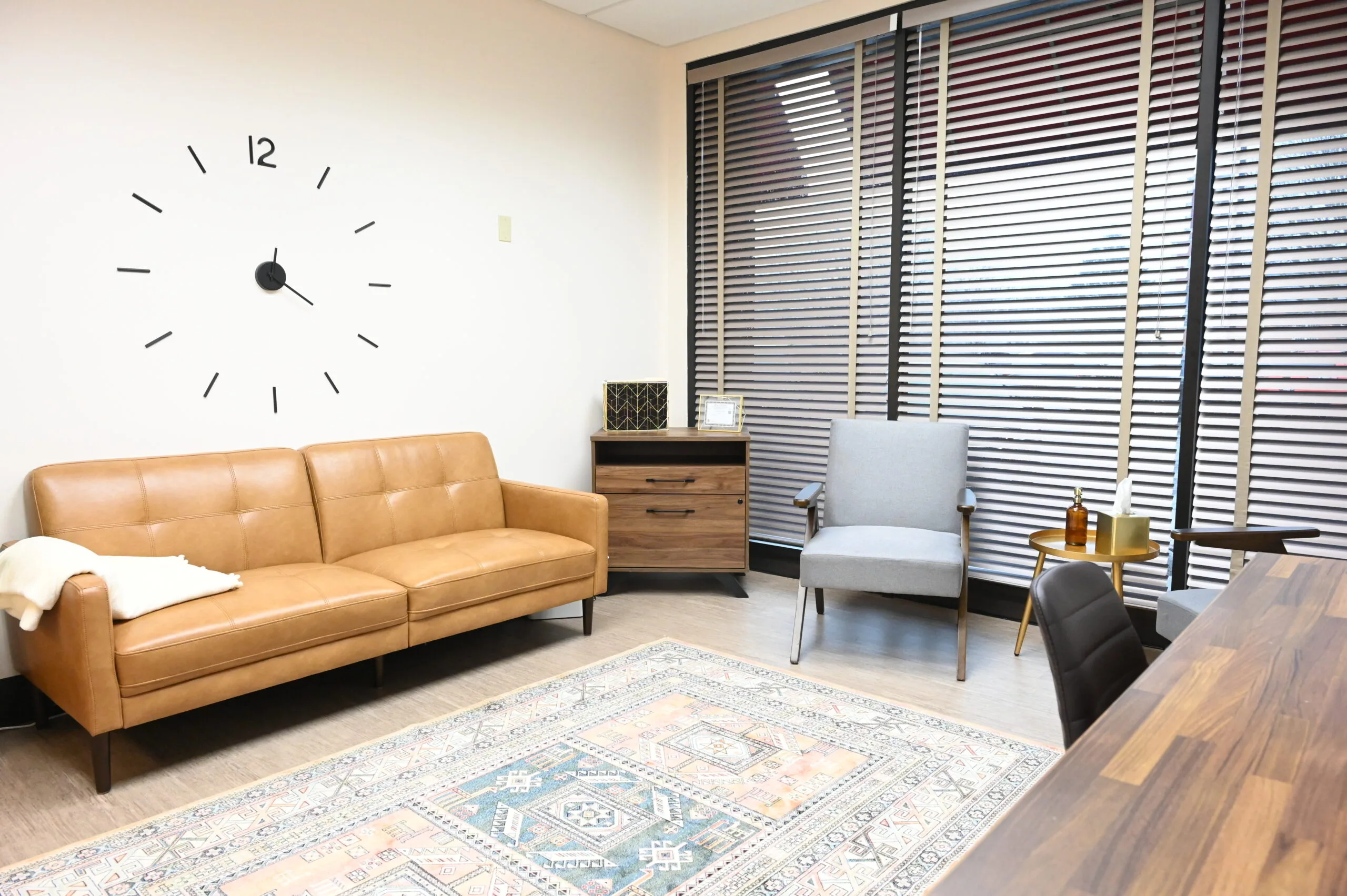 Modern waiting room with tan leather sofa, gray armchair, wooden desk, wall clock, and louvered window blinds