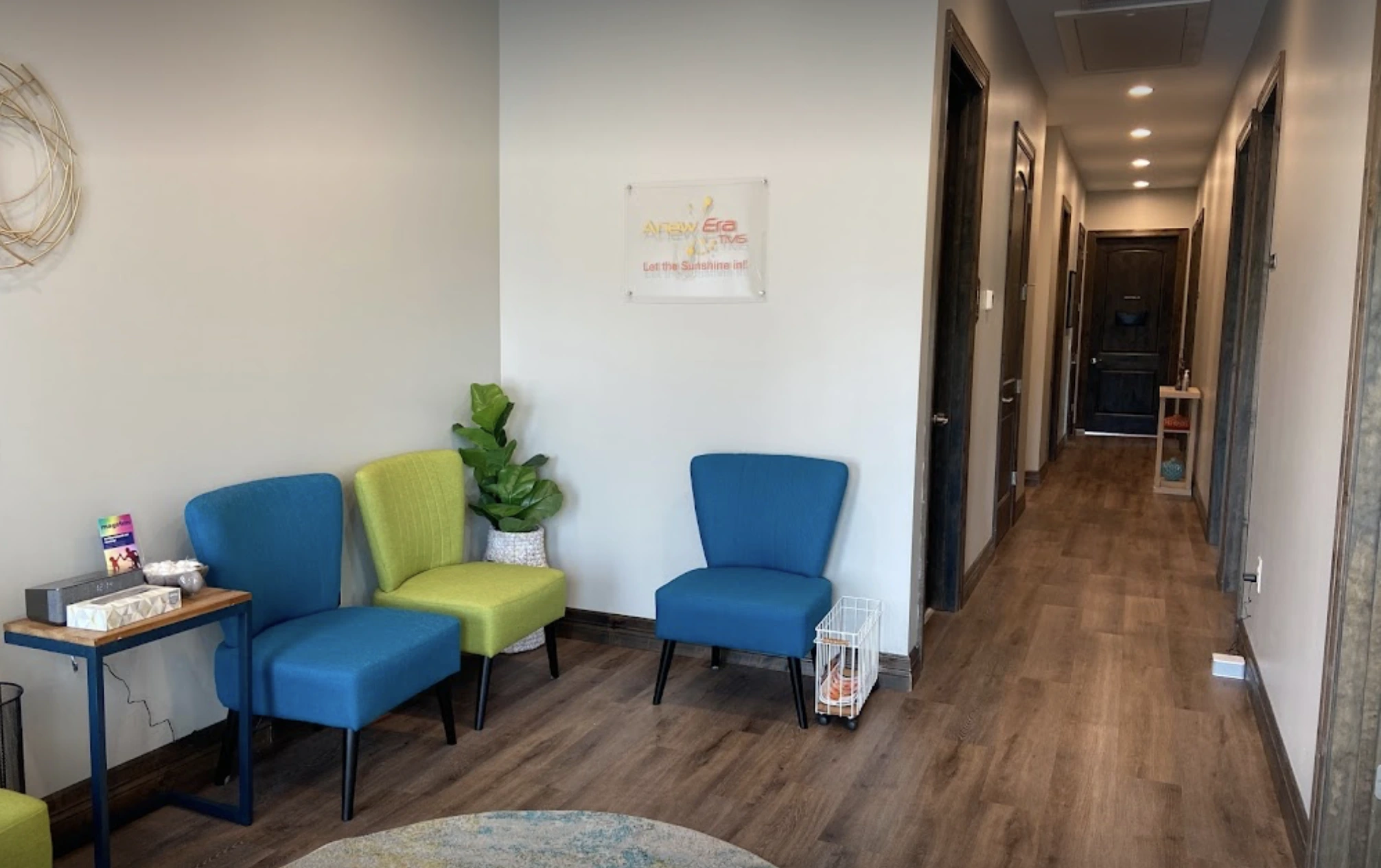 Hallway with blue and green chairs, potted plant, small side table, and treatment room doors leading to clinical areas