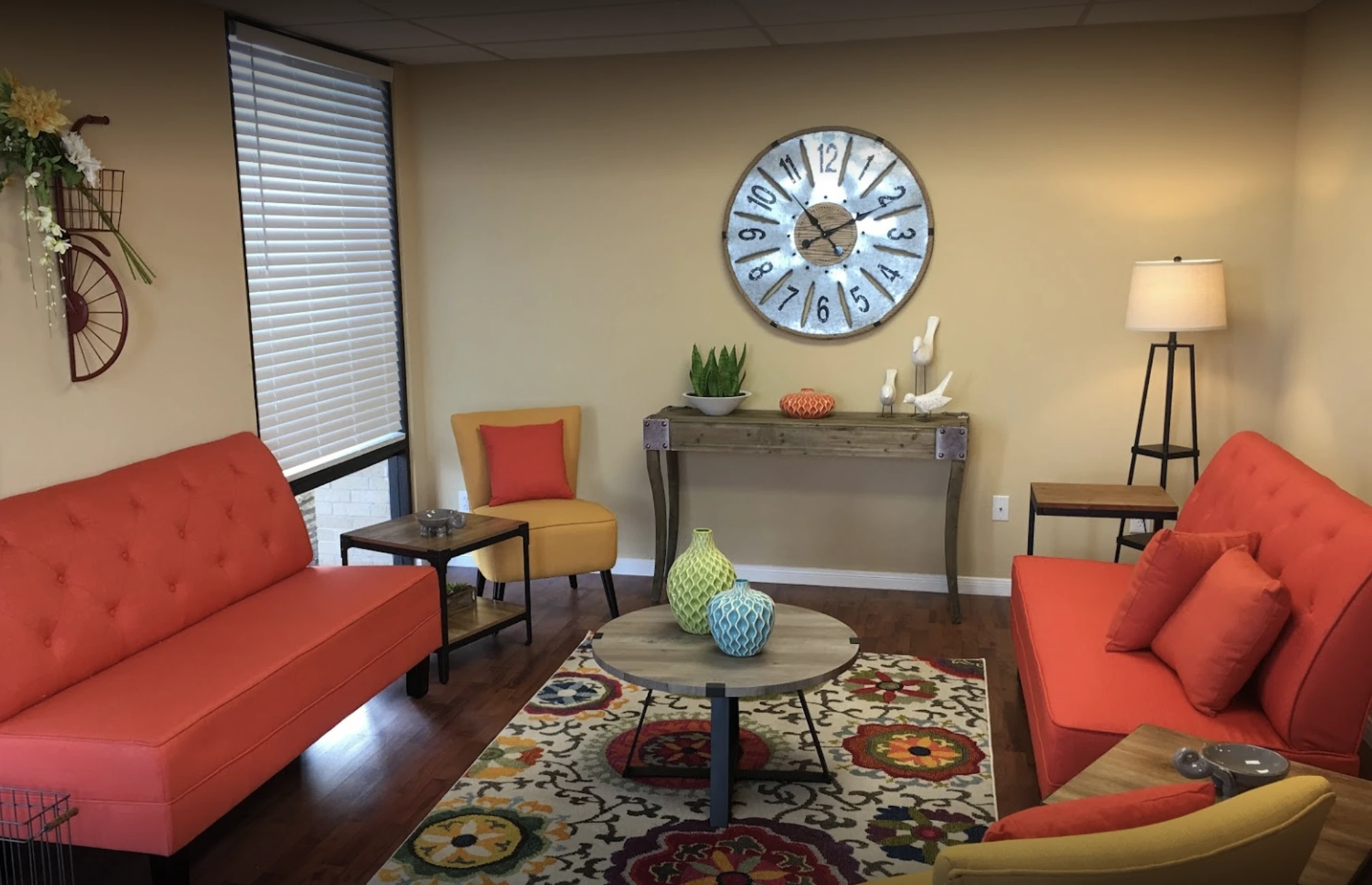 Welcoming waiting room with red sofas, colorful patterned rug, round blue clock, and potted plant on console table