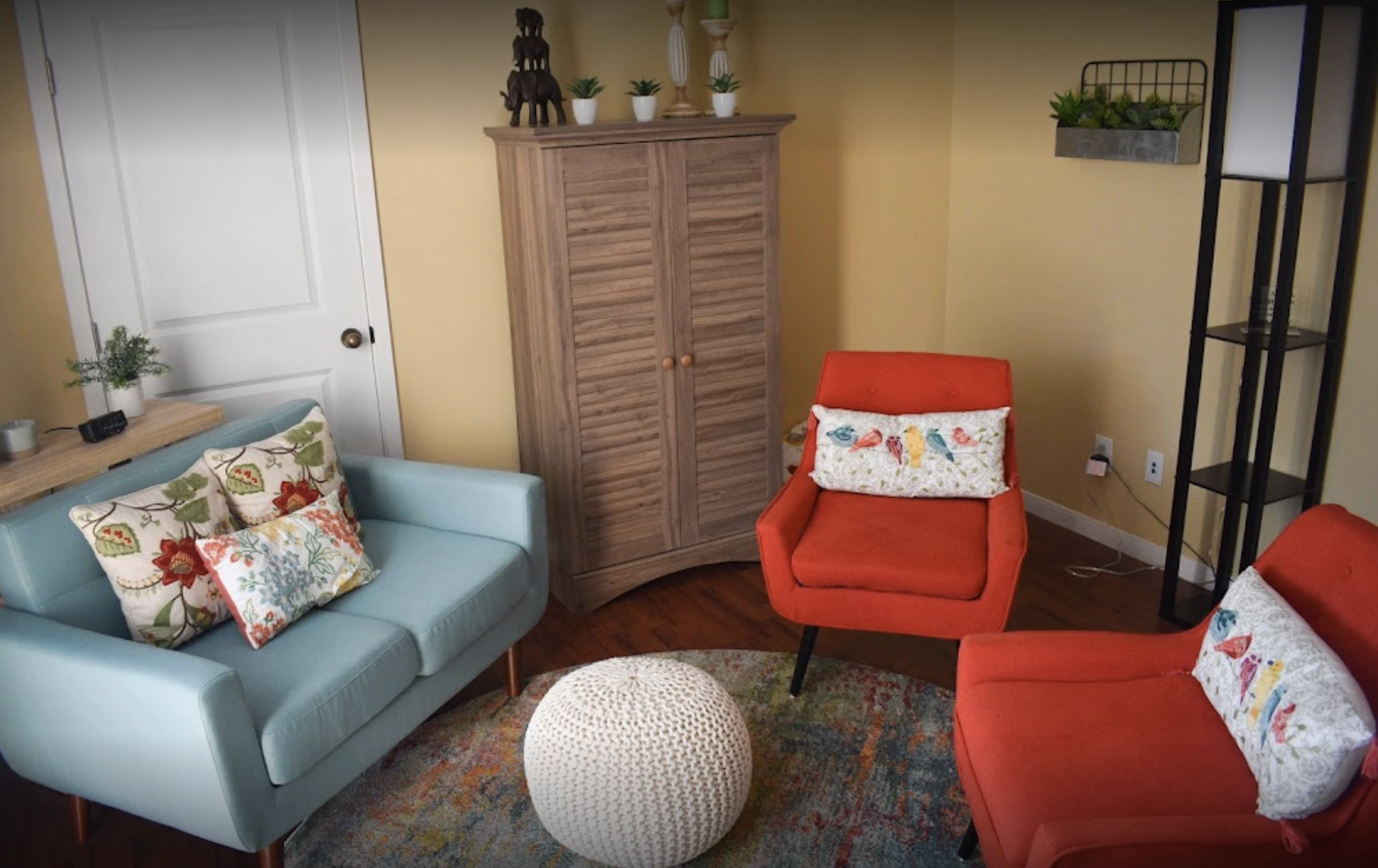 Waiting area with light blue sofa, orange chairs, wooden storage cabinet, white pouf, and colorful throw pillows