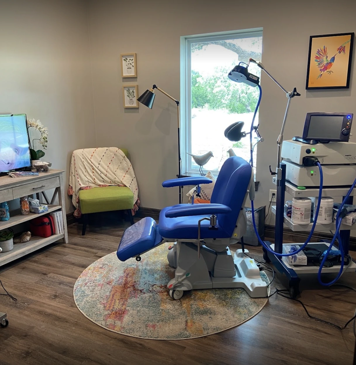 TMS treatment room with blue patient chair, overhead stimulation device with dual lighting arms, desk, and glass door with outdoor view