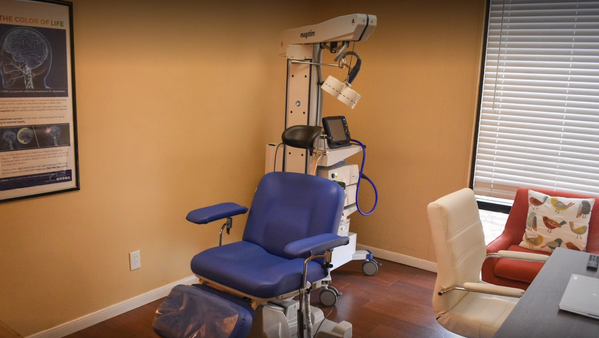 TMS treatment room with orange walls, blue patient chair, overhead magnetic stimulation device, and anatomical poster