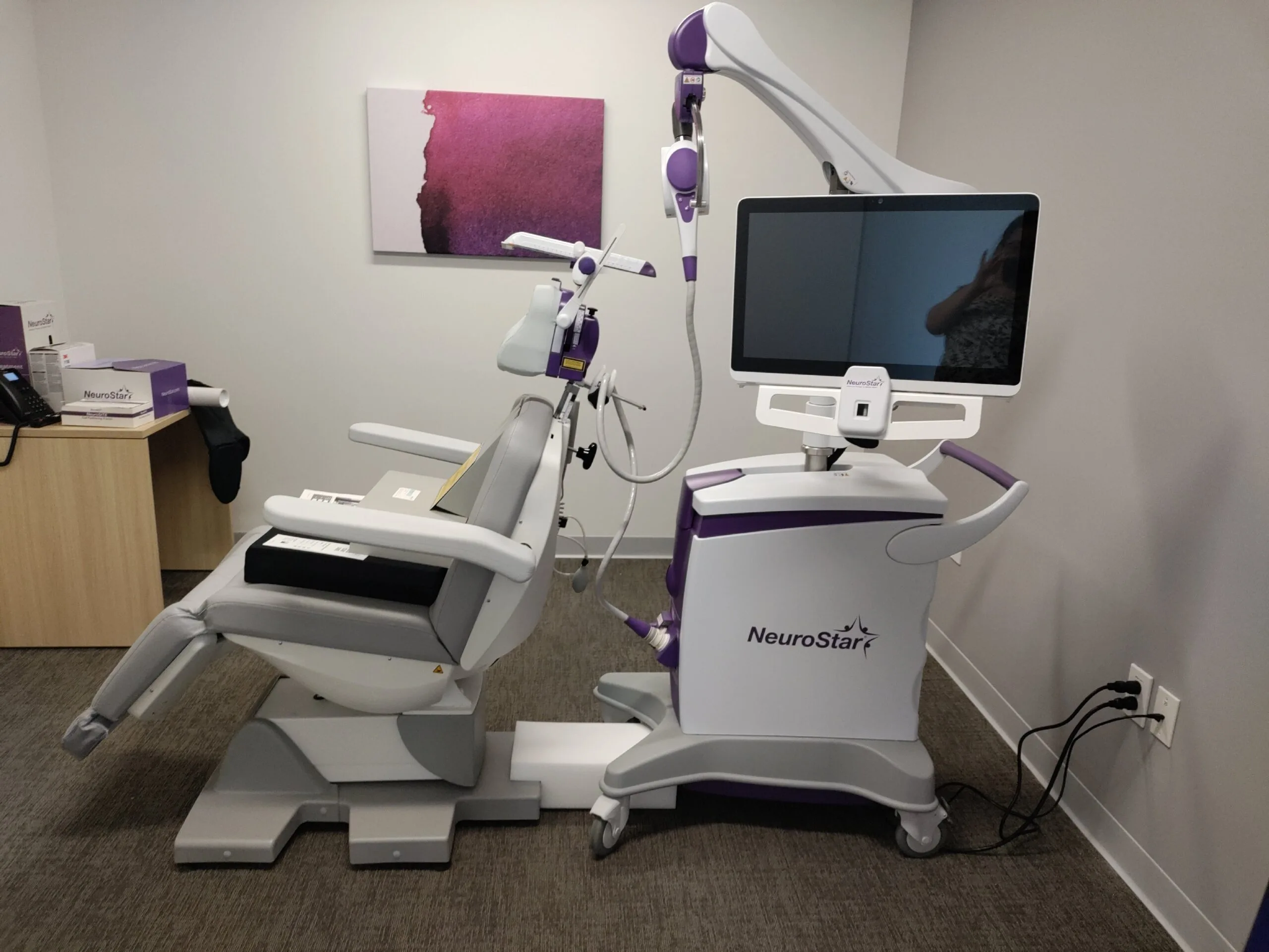NeuroStar TMS treatment chair with white and purple device, monitor, and coil positioned above patient seating area