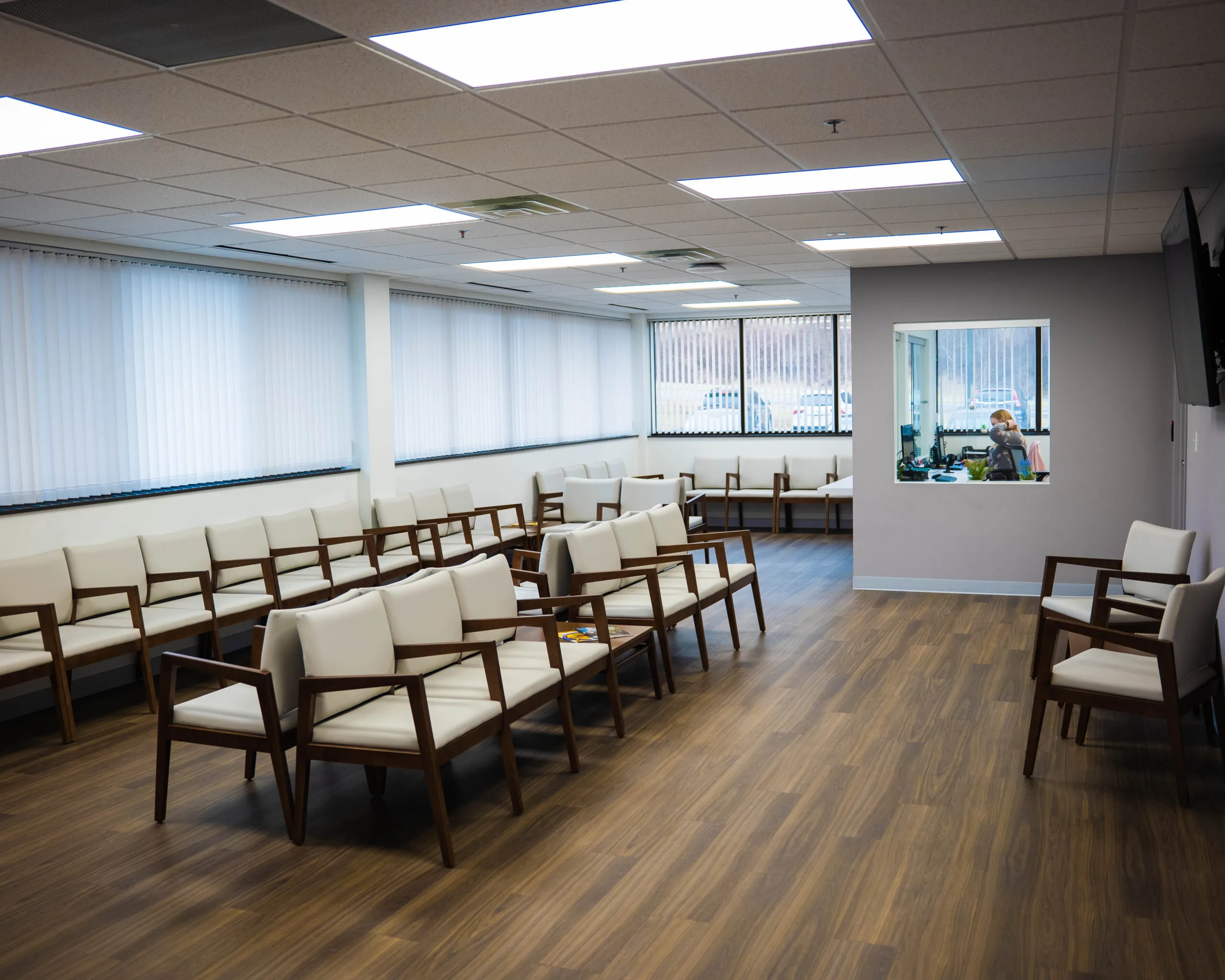 Large waiting room with rows of cushioned wooden chairs, windows with blinds, and interior window view