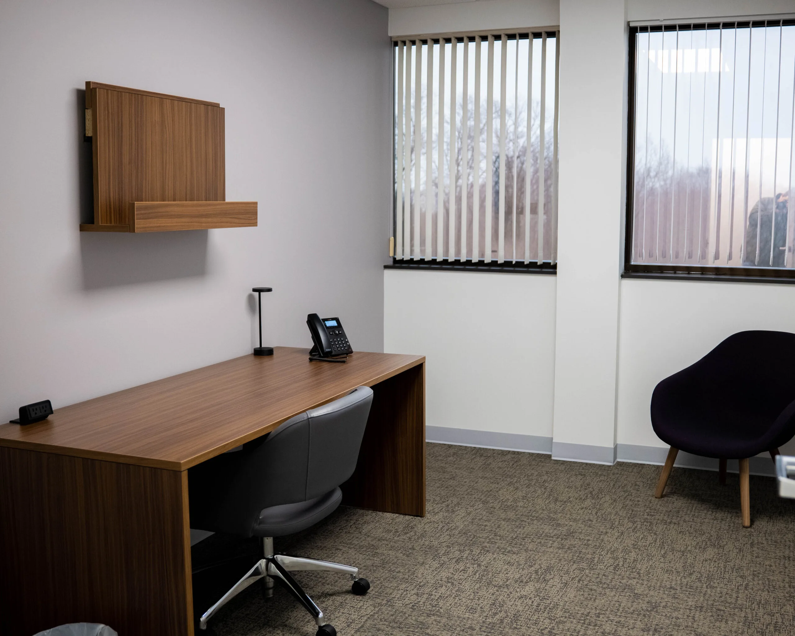 Consultation office with wooden desk, black ergonomic chair, and window blinds