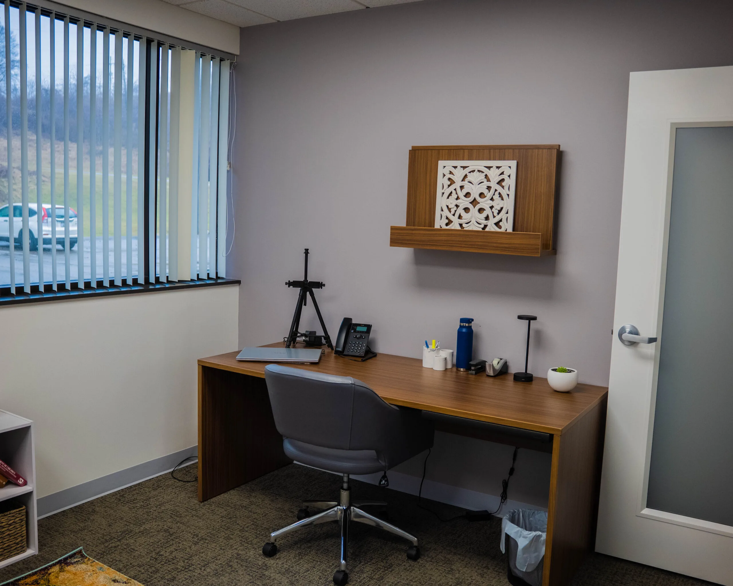 Office workspace with wooden desk, black chair, desk phone, and framed wooden wall art with vertical blinds