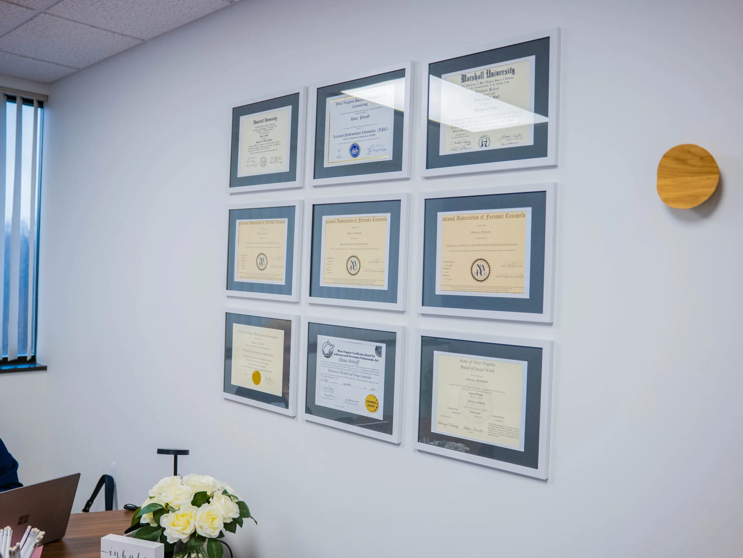 Nine framed certificates and diplomas displayed on wall above desk with white flowers and wooden accents
