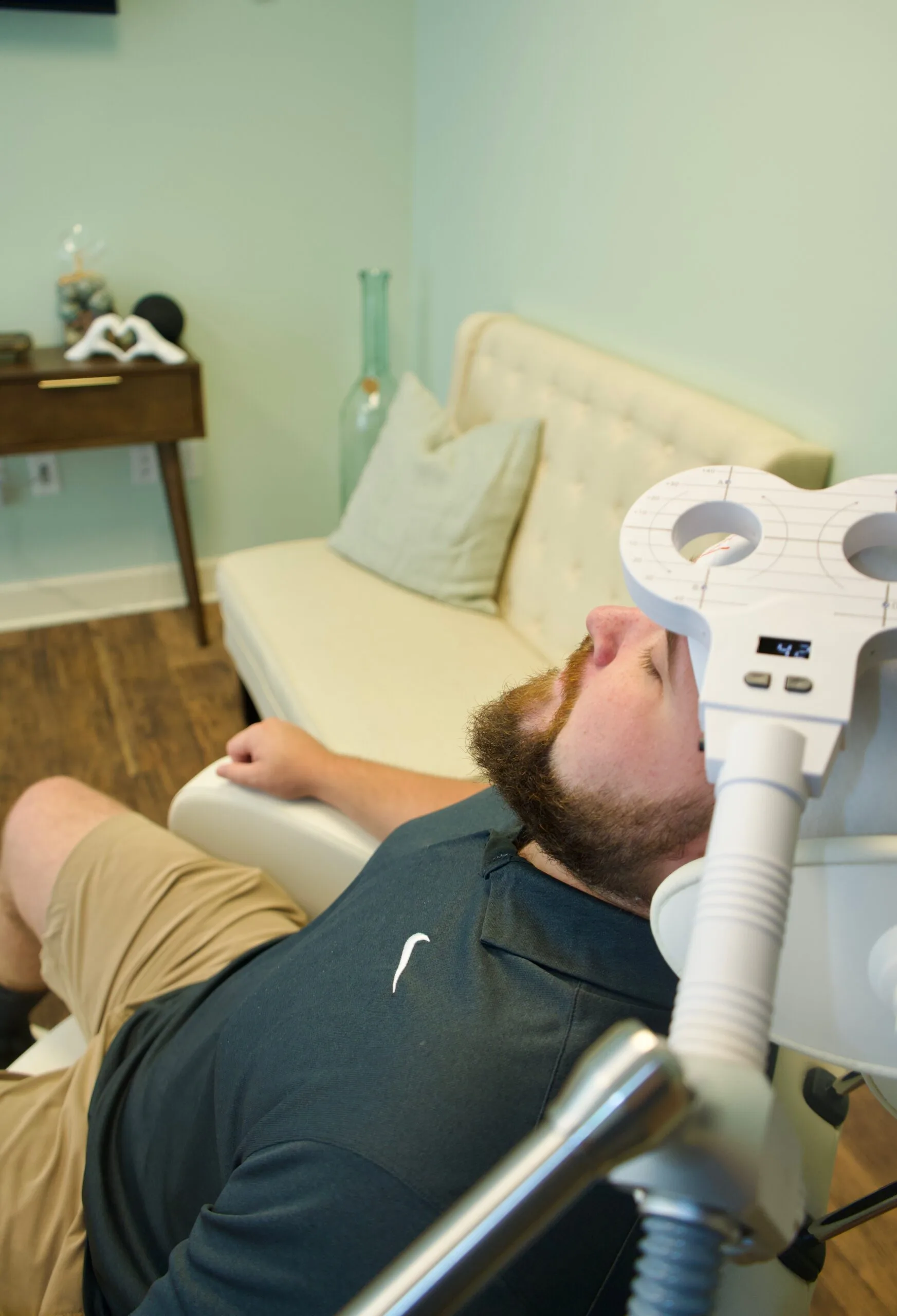 Patient receiving TMS therapy with magnetic coil device positioned against head while seated in treatment room