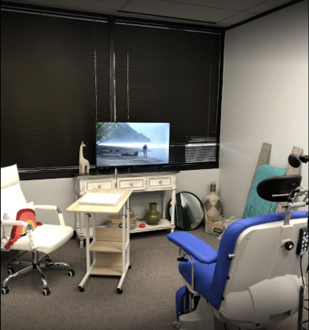 TMS treatment room with blue and white patient chair, TV displaying nature scene, whitewashed furniture, and medical equipment