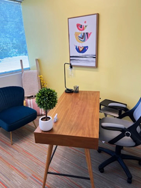 TMS therapy office with yellow walls, wooden desk, colorful abstract artwork, green chair, and window