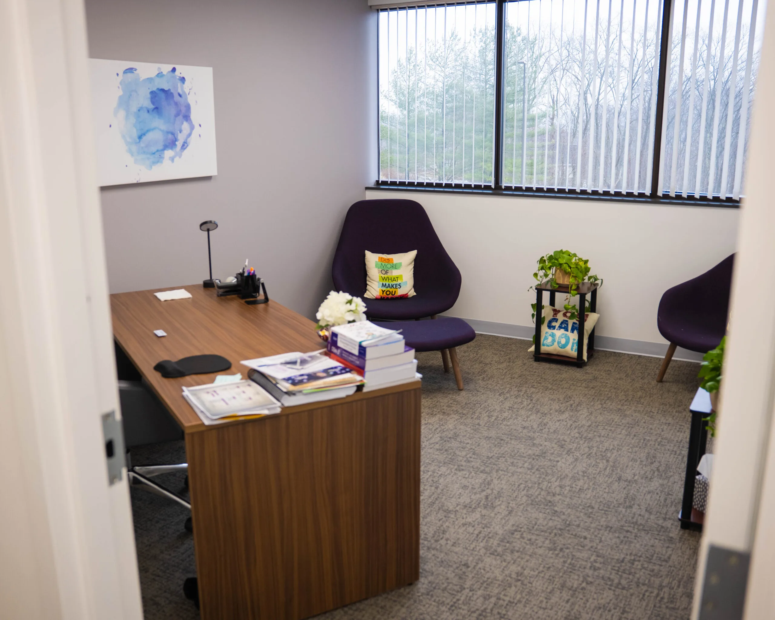Consultation room with wooden desk, black accent chair, blue abstract wall art, and potted plants by windows