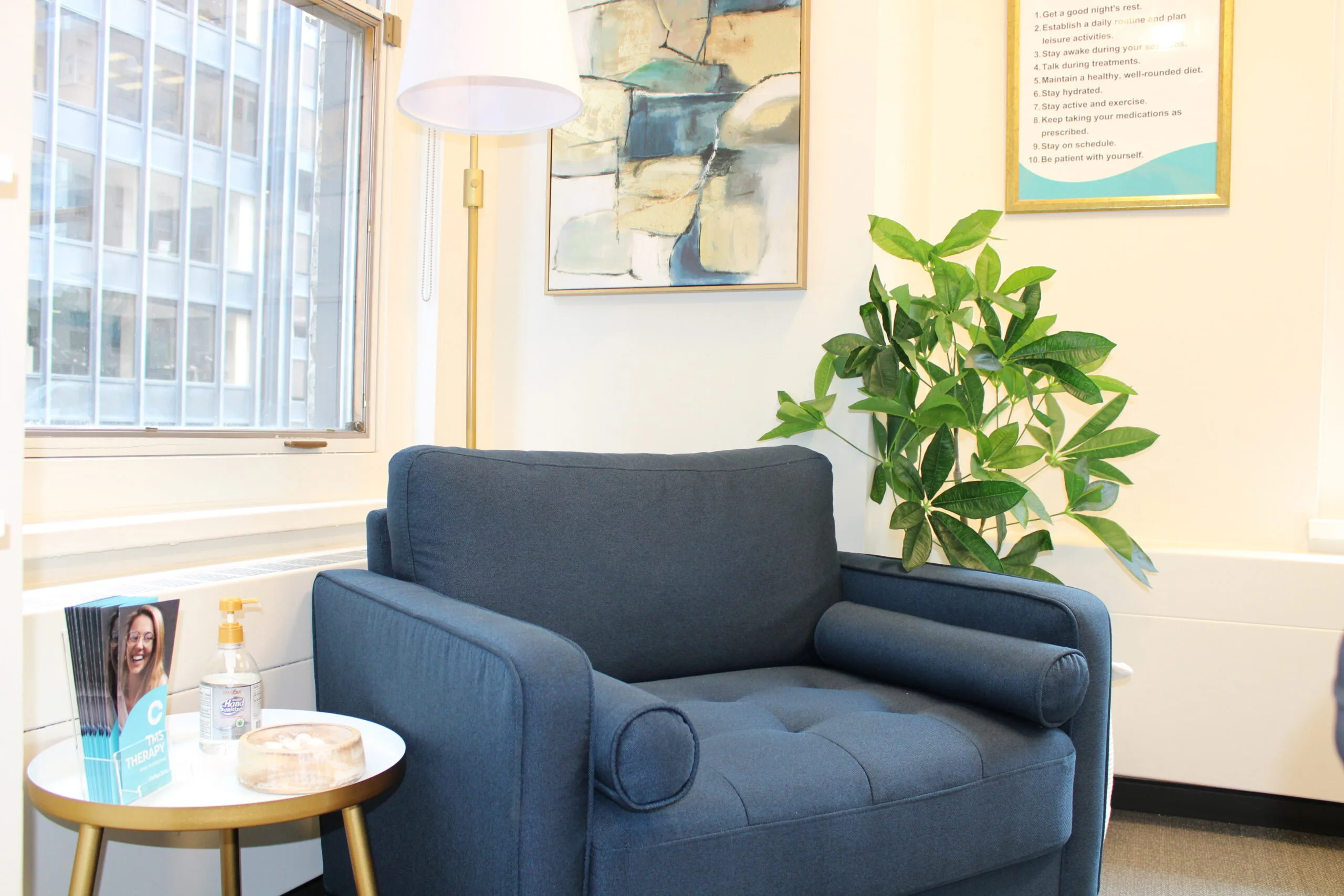Waiting room with dark blue armchair, small round side table, potted plant, and framed artwork