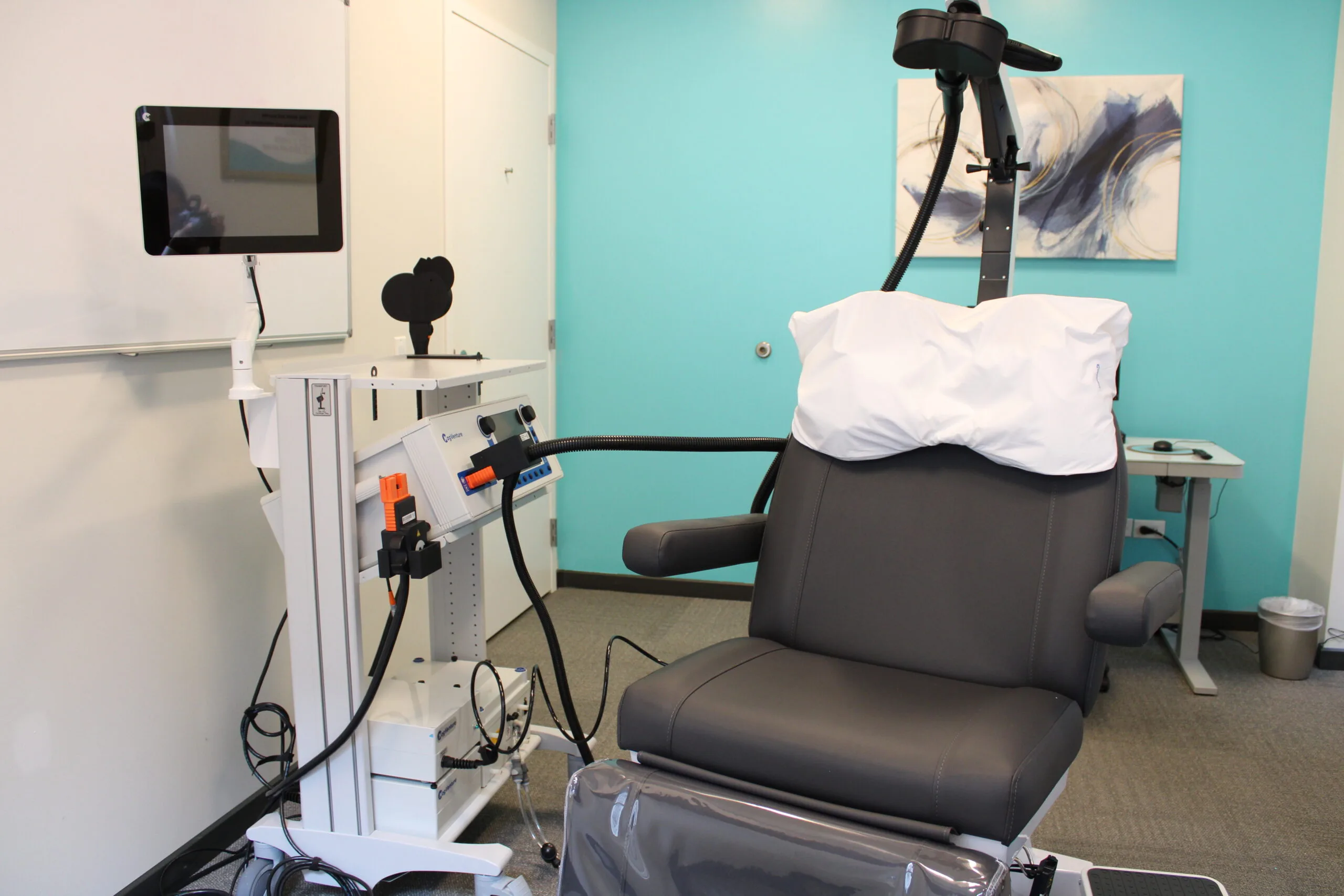 TMS therapy treatment room with patient chair, white medical equipment with orange accents, monitor, and coil applicator mounted above