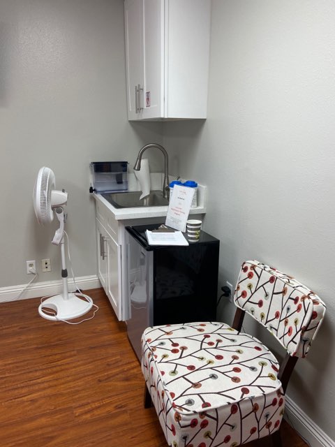 Kitchenette with sink, white cabinets, small fan, and patterned seating chair on wooden floor