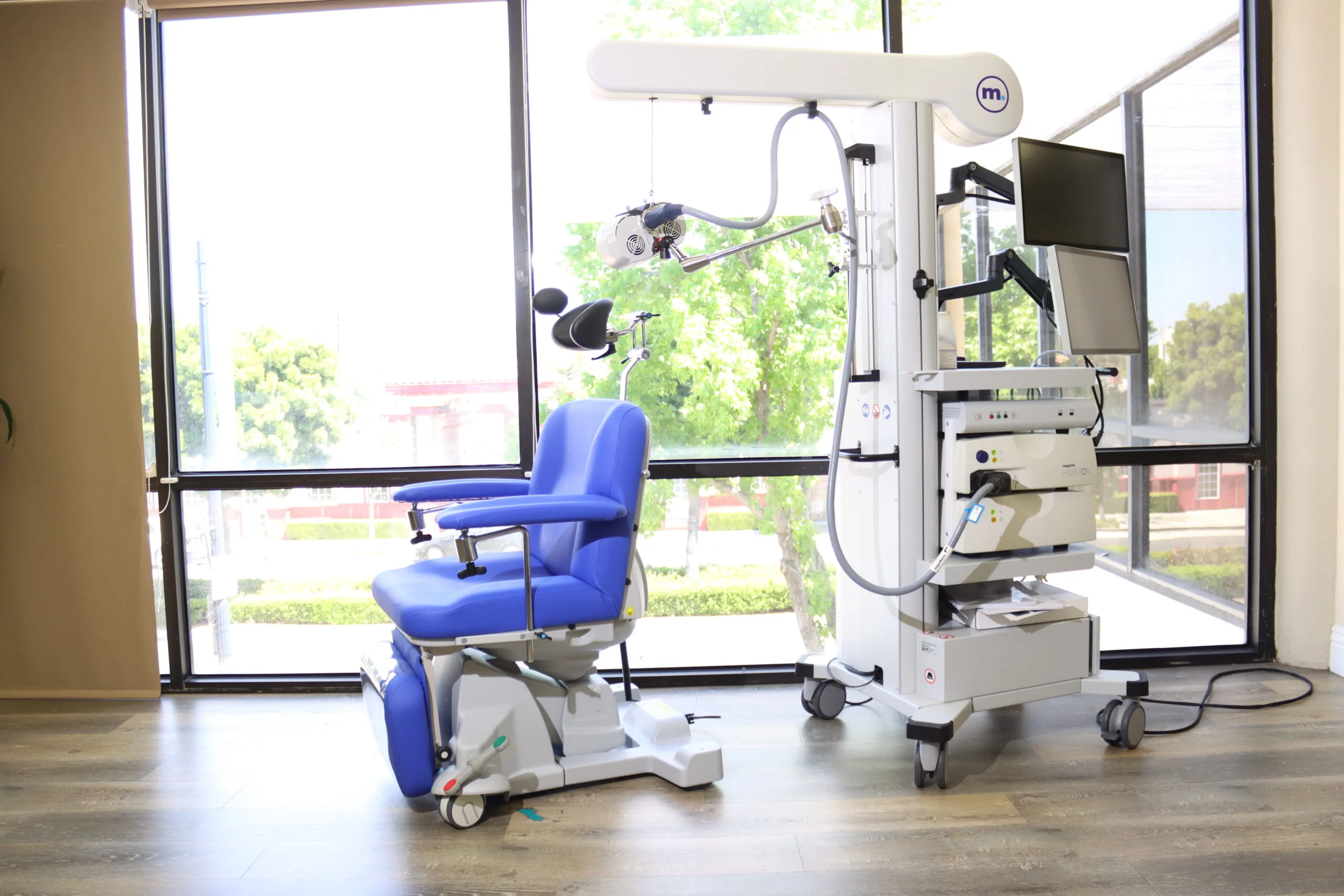 TMS therapy room with blue treatment chair, overhead magnetic coil equipment, and natural light from windows