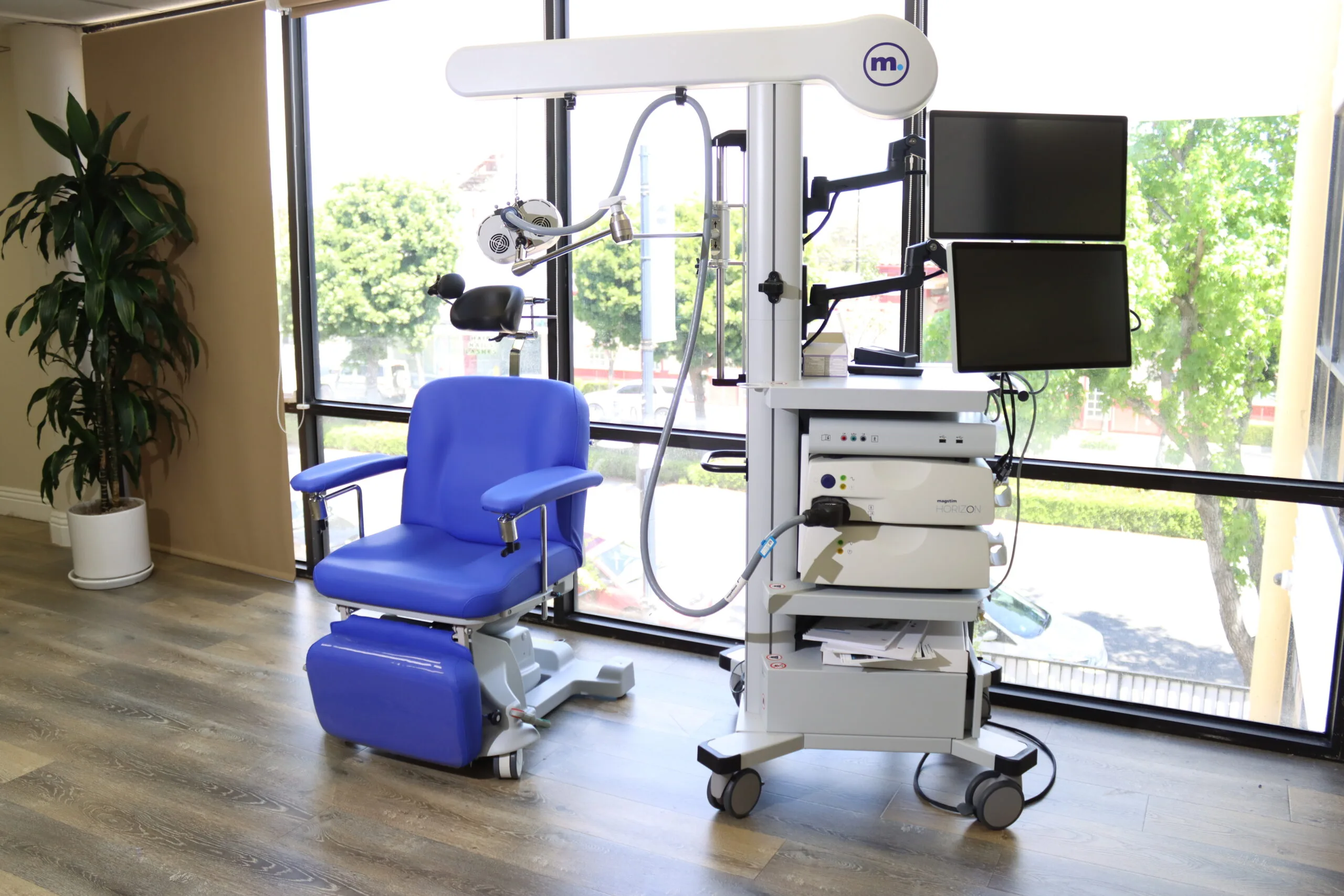 TMS treatment room with blue patient chair, medical equipment on adjustable arm, and large windows