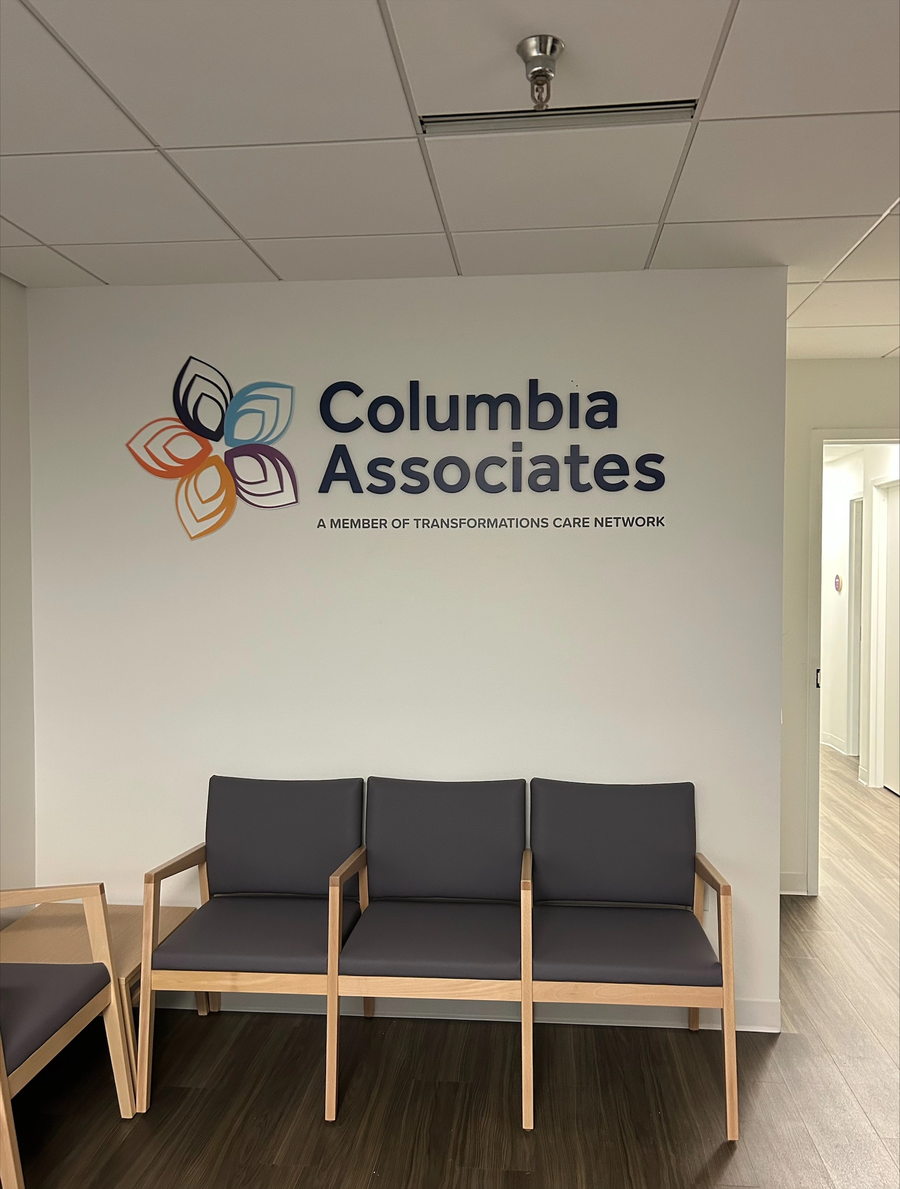 Columbia Associates waiting room with three-seat wooden chair set and wall signage reading "Columbia Associates, A Member of Transformations Care Network"