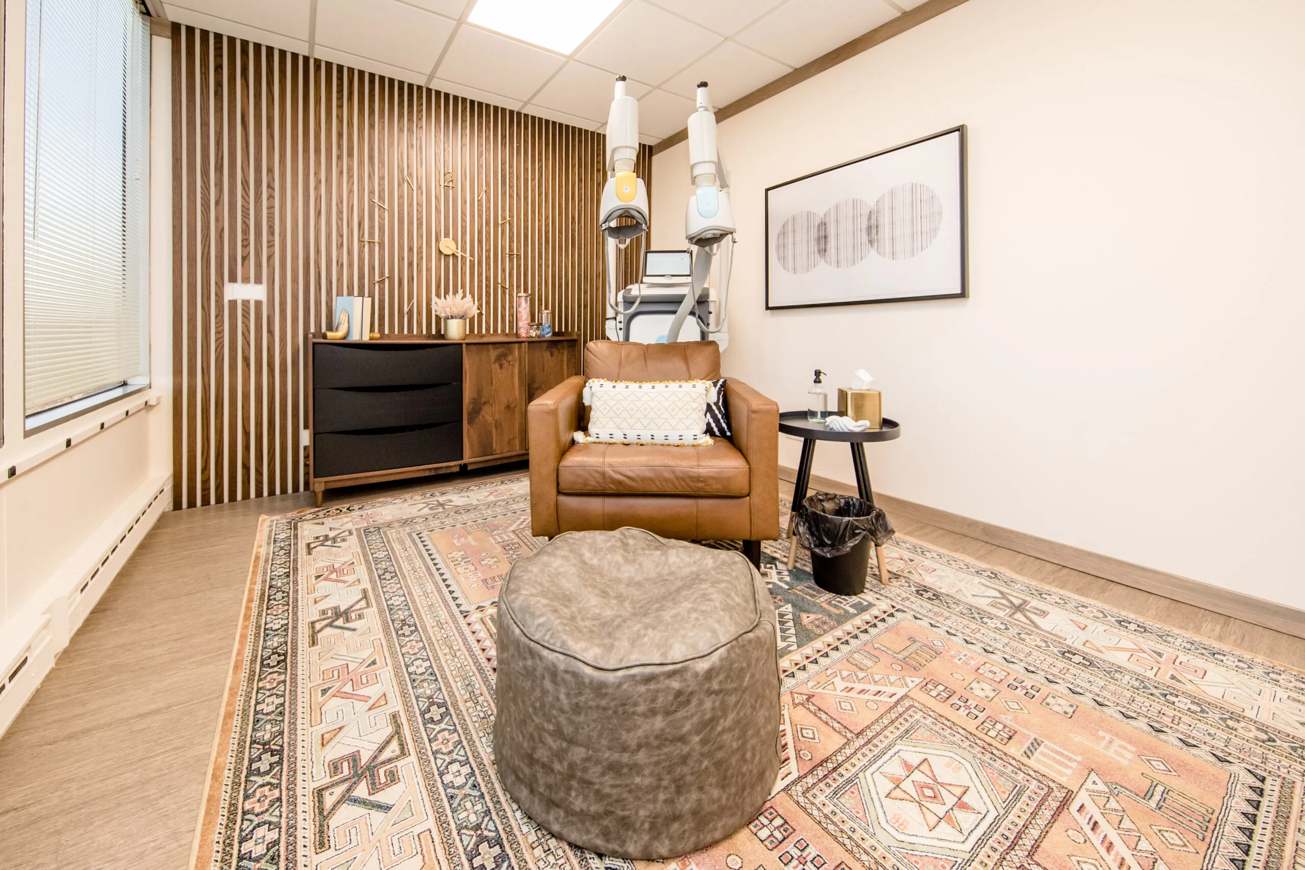 TMS therapy treatment room with brown leather armchair, gray ottoman, wooden furniture, and ceiling-mounted medical equipment
