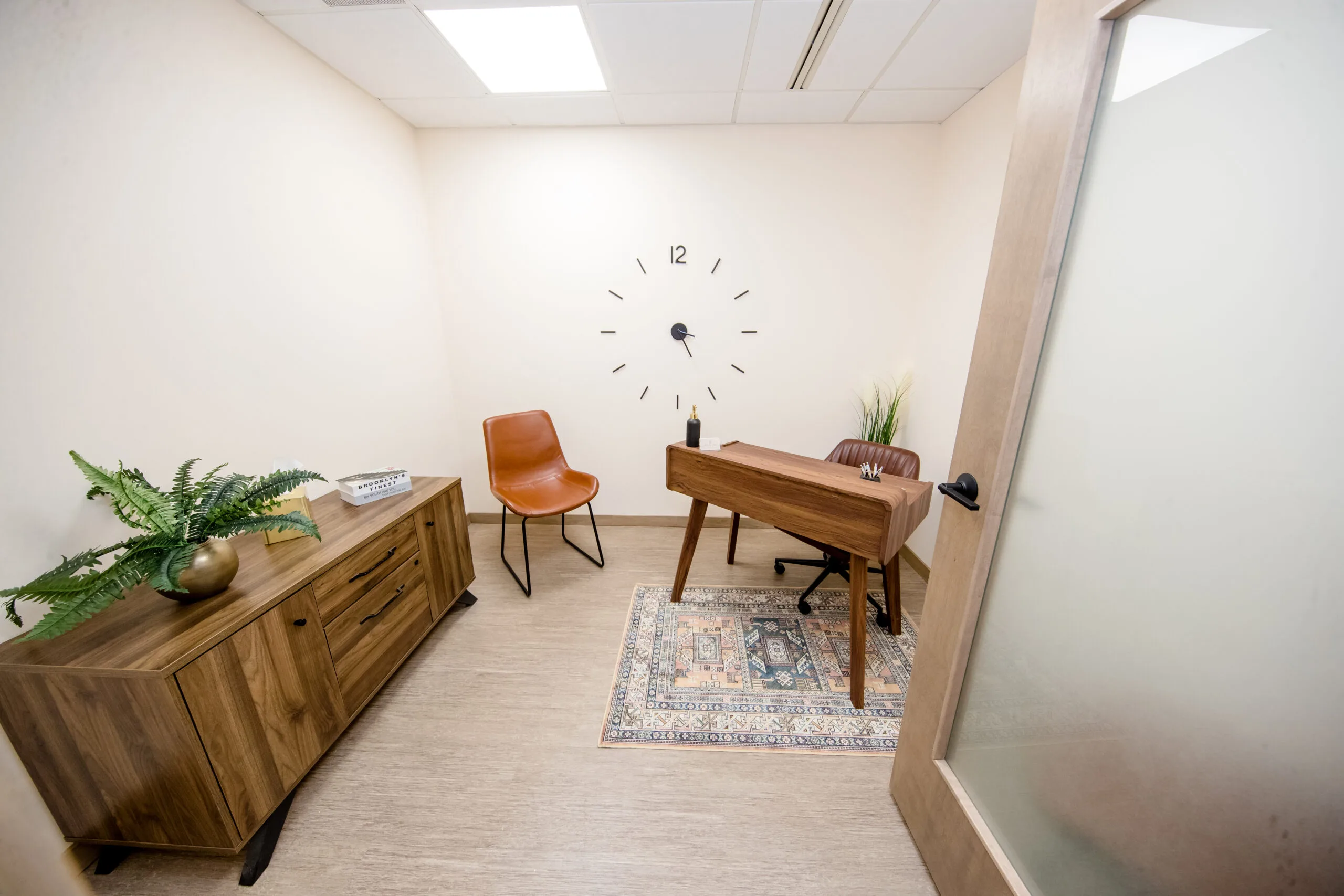 Waiting area with wooden dresser, orange chair, wall clock, potted plants, and wooden desk