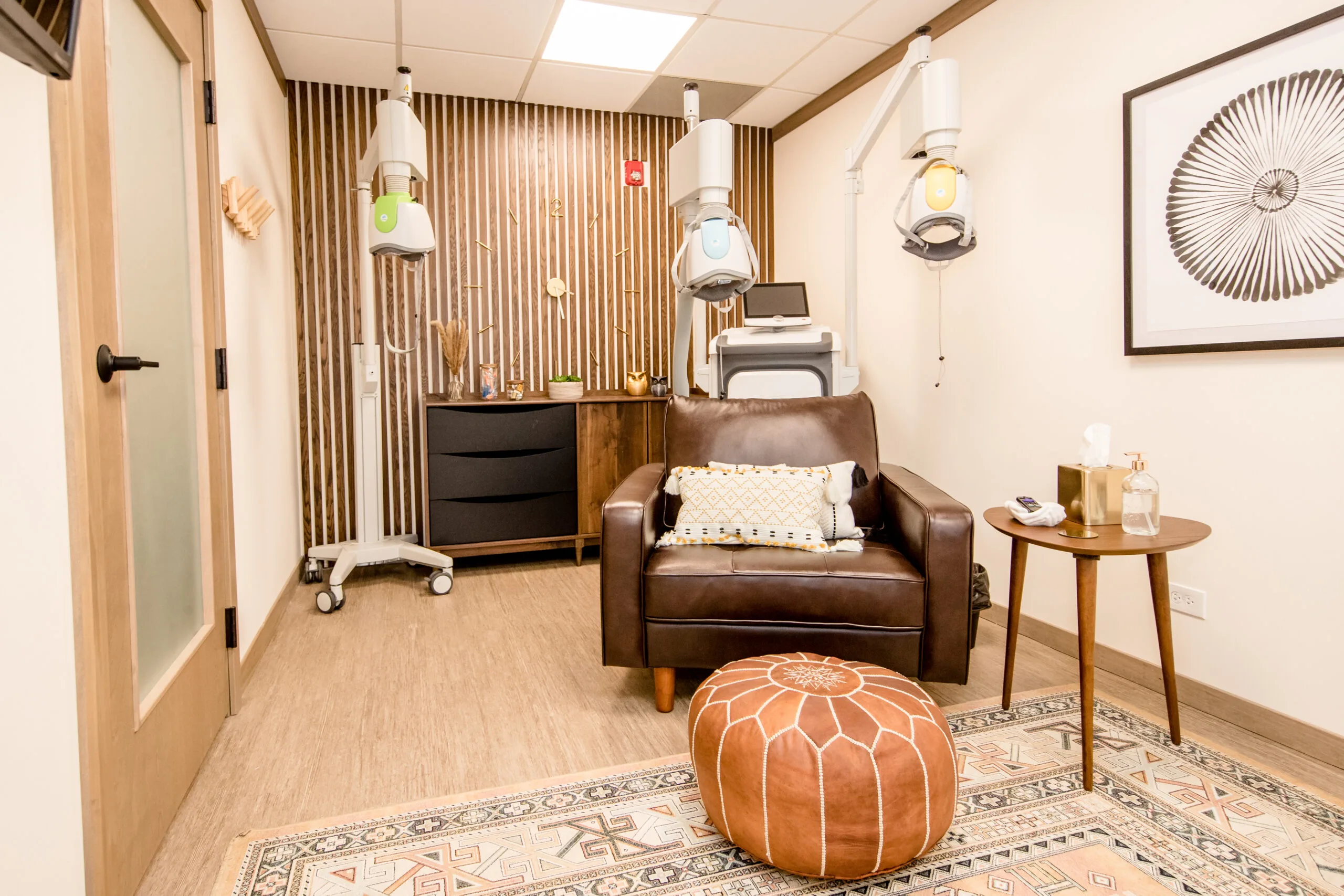 TMS treatment room with brown leather chair, orange ottoman, pendant lights, and ceiling-mounted therapy equipment