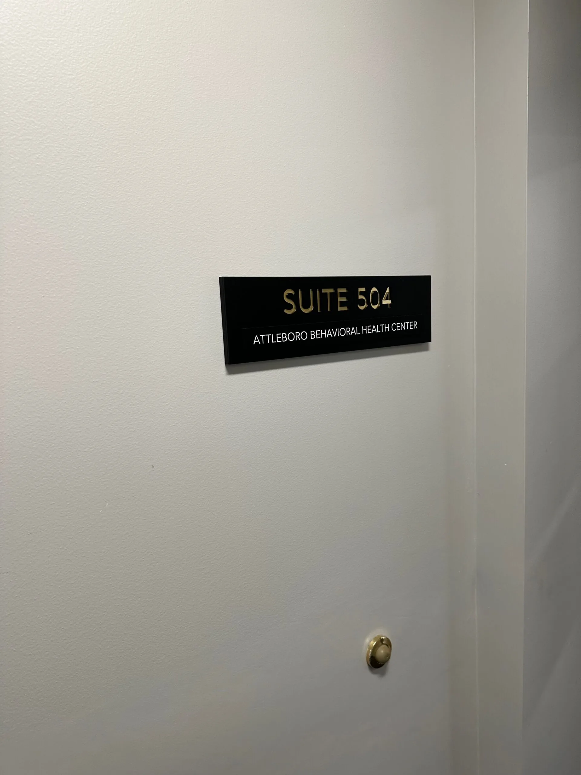 Black plaque on white wall reading 'SUITE 504 ATTLEBORO BEHAVIORAL HEALTH CENTER'