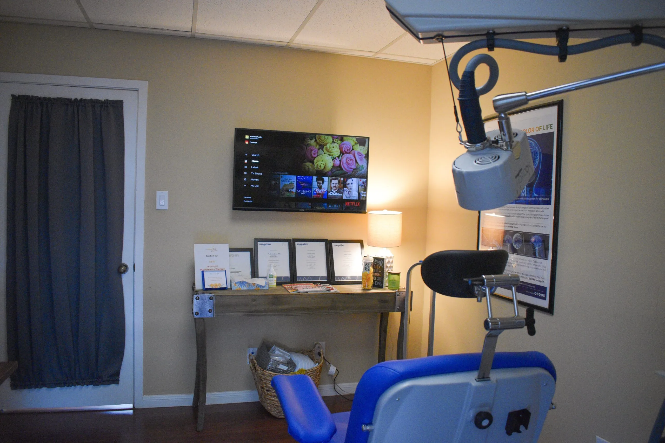 TMS treatment room with blue patient chair, mounted coil device, monitor displaying media content, and treatment information materials on wall
