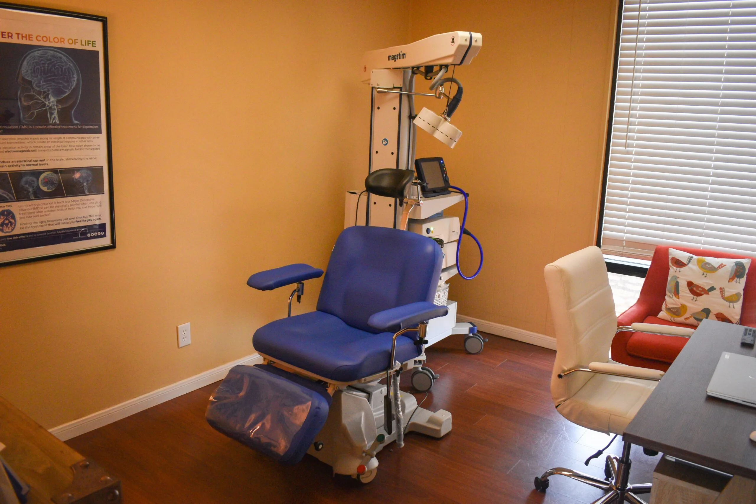 TMS treatment room with blue patient chair, overhead magnetic coil device, brain imaging poster, and seating area with red pillows