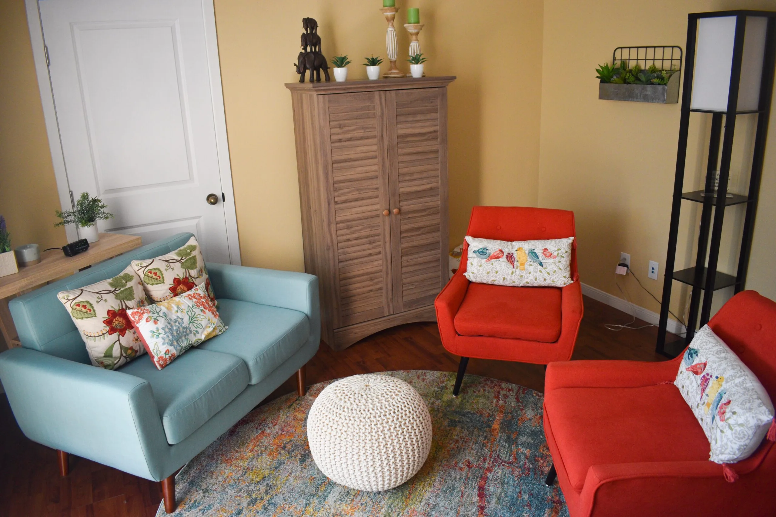 Therapy waiting room with turquoise and red chairs, white pouf, wooden cabinet, plants, and floral throw pillows