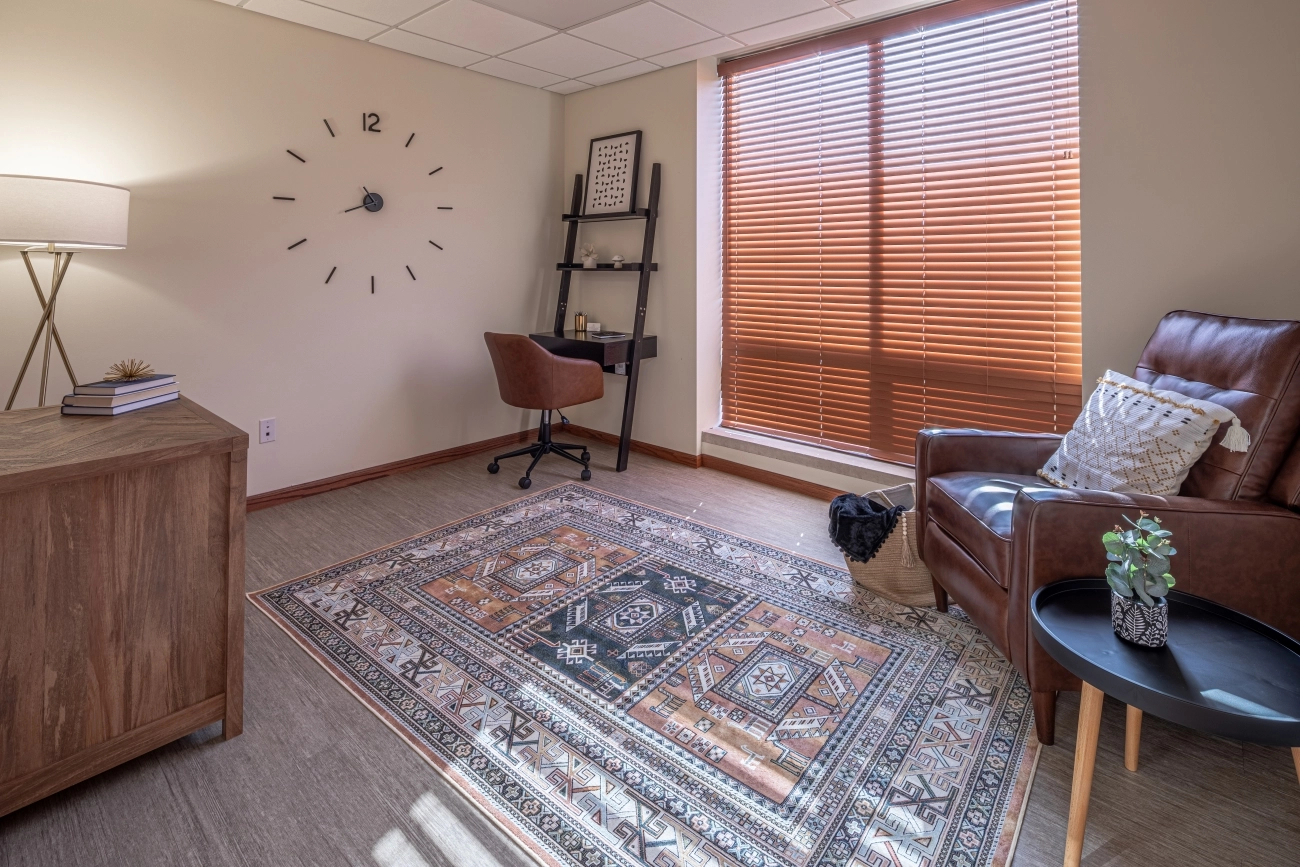 Private therapy room with brown leather chair, desk with lamp, shelving unit, and large window with blinds