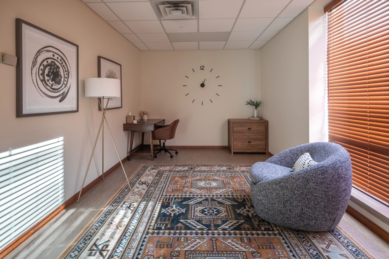 Therapy office with gray armchair, wooden desk, framed artwork, wall clock, and orange blinds on windows